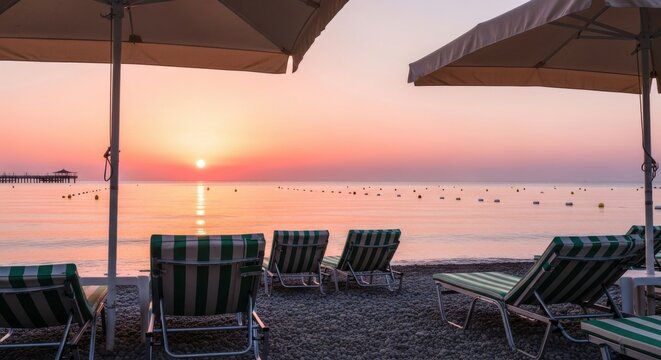 Serene sunrise over the sea viewed from a pebble beach with several empty striped lounge chairs and large umbrellas providing shade in the foreground - Powered by Adobe