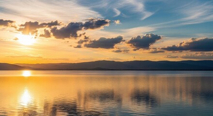 Breathtaking sunset over a calm lake with vibrant orange and yellow reflections stretching across the water surface under a dramatic, cloudfilled sky with distant mountains silhouetted on the horizon