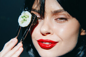 A young woman with striking red lips enjoys sushi, holding a piece with chopsticks, against a dark, blurred background, exuding confidence and allure.