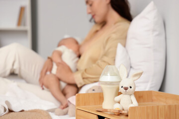 Bottle of milk with toy on table against woman breastfeeding her baby in bedroom, closeup