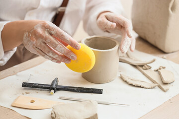 Young female potter shaping clay pot with sponge at table in home studio, closeup