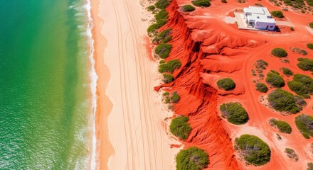 Aerial view of vibrant red earth cliff meeting a pristine sandy beach alongside turquoise ocean water, featuring a solitary white building perched on the dramatic coastline