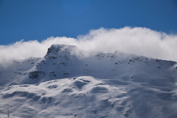 Snow-covered alpine mountains with dramatic clouds rolling over the ridge. Atmospheric winter landscape with soft light, rugged terrain, and high-altitude scenery.