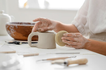 Obraz premium Young female potter shaping clay cup with pottery sponge at table in home studio, closeup