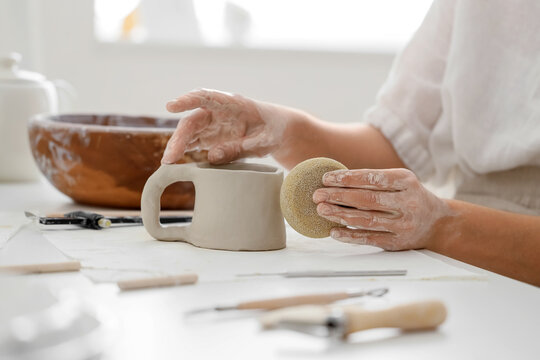 Young female potter shaping clay cup with pottery sponge at table in home studio, closeup