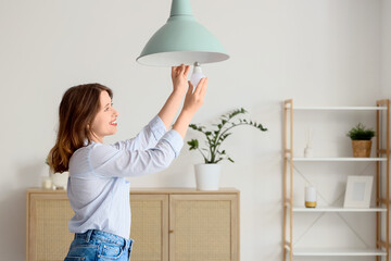 Pretty young woman changing light bulb of hanging lamp at home