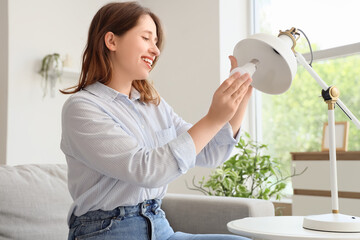 Pretty young woman changing light bulb of desk lamp in living room