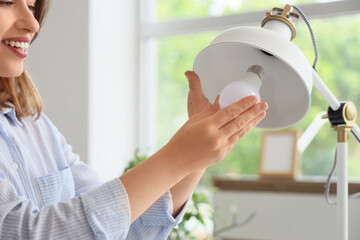 Pretty young woman changing light bulb of desk lamp at home, closeup