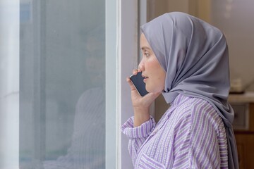 A young Asian Muslim woman standing by the window while talking on the phone