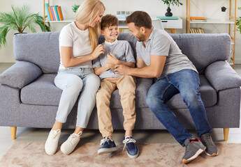 Happy family at home, cheerful excited parents tickling beloved kid son, playing with child on sofa in modern home interior, sitting laughing, smiling for joy, enjoying family leisure weekend