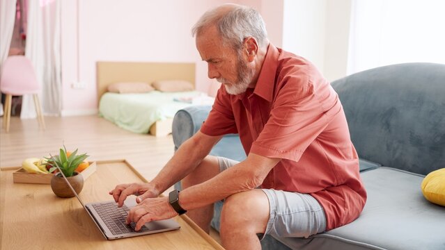 Senior man using a laptop on a coffee table, seated on a couch in a comfortable living room with soft lighting and modern decor - Powered by Adobe
