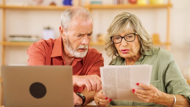 Senior couple attentively reviewing paperwork together, using a laptop for assistance. They appear focused and engaged in a home office setting