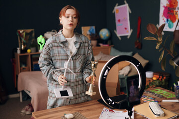 Caucasian teenage girl recording video in bedroom holding wind turbine model and trophy, standing behind desk with smartphone on tripod, engaging in educational or creative activity