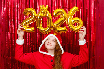 Cute girl in a santa costume holding golden balloons with the shape 2026 on a red background.