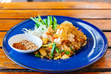 Authentic Pad Thai with shrimp, topped with crispy shredded egg and served with crushed peanuts and chili flakes on a blue plate over a rustic wooden table. Famous Thai street food and Asian cuisine.