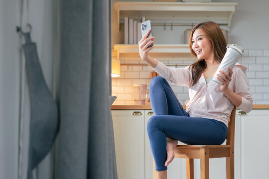 A beautiful young woman relaxes in her living room after an intense home workout, sipping refreshing water while video calling friends on her phone and capturing a glowing post-yoga selfie share life