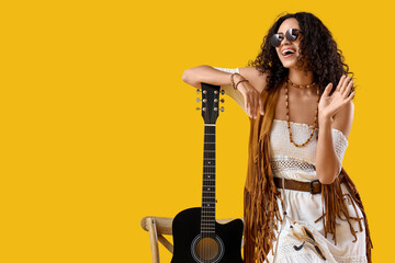 Happy young African-American woman with acoustic guitar waving on yellow background