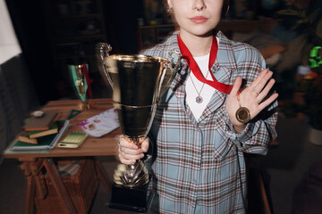 Caucasian teenage girl holding large trophy in one hand and showing gold medal with other hand, standing in room with study materials visible in background, face partially visible