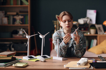 Caucasian teenage girl assembling wind turbine model at desk, focusing on renewable energy project, surrounded by study materials and miniature wind turbines in bedroom or study room
