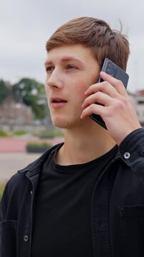 Young man with brown hair and a black jacket pushes talk and starts a conversation on his smartphone, standing outdoors in a city environment surrounded by daylight, modern buildings, and greenery.