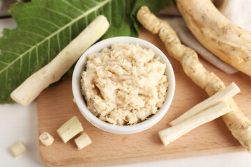 Bowl of horseradish sauce with roots, slices and leaves on white wooden background