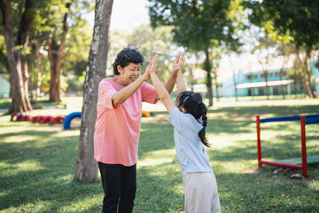 Fototapeta premium Joyful moment between grandmother and granddaughter in a sunny park engaging in playful activities, surrounded by green trees and vibrant playground equipment.