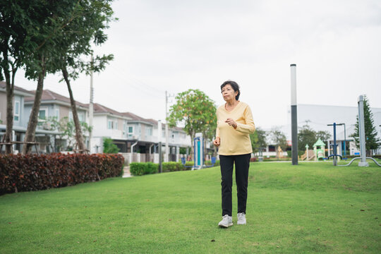 Active senior woman walking confidently in a park setting surrounded by greenery and modern residential buildings under a cloudy sky