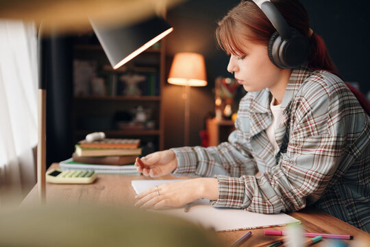 Caucasian teenage girl wearing headphones studying at desk, drawing in notebook with colored pencils and calculator nearby, focused on homework or creative project in bedroom - Powered by Adobe