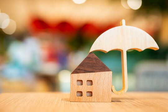 Wooden house model protected by a wooden umbrella on a wooden table with bokeh background, symbolizing home insurance, safety, real estate protection, and financial security concept.