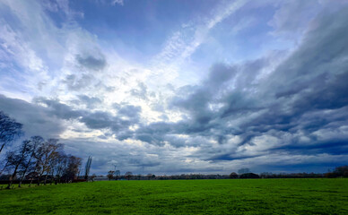 A stunning view of a dynamic sky filled with clouds above a vibrant green field with colorful...