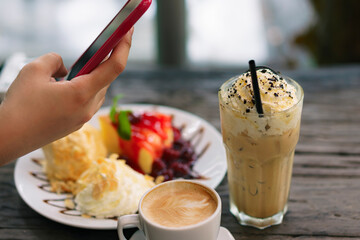 Hand holding smartphone taking photo of iced coffee, hot latte, and dessert at cozy café table, representing social media food trends, modern cafe lifestyle, and digital dining culture.