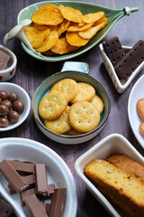Assorted party snacks on table featuring chocolate cream sandwich biscuits, sugar coated heart cookies, wafer chocolates, chocolate coated caramel balls, salted crackers, and crispy potato chips.