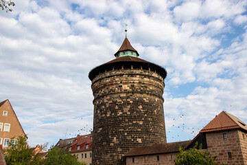 Historic stone tower with a conical roof stands tall against a blue sky filled with clouds, surrounded by traditional buildings and greenery, showcasing architectural heritage and urban landscape