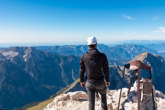 A mountaineer stands near the summit bell of Jof di Montasio, gazing at the endless layers of the Julian Alps and Carnic Alps, celebrating the achievement of reaching the peak in Friuli, Italy.