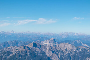 Majestic limestone peaks of the Julian Alps rise dramatically under a clear blue sky, overlooking the distant Dolomites ranges from the summit of Jof di Montasio in Friuli Venezia Giulia, Italy.