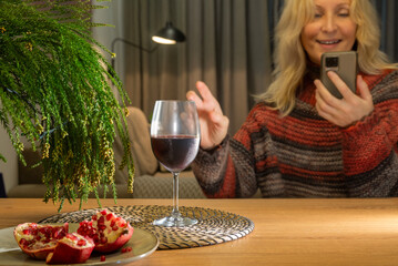 A glass of red wine, pomegranate and green fir branches on a wooden table against a background of a talking on the phone happy woman. Close-up