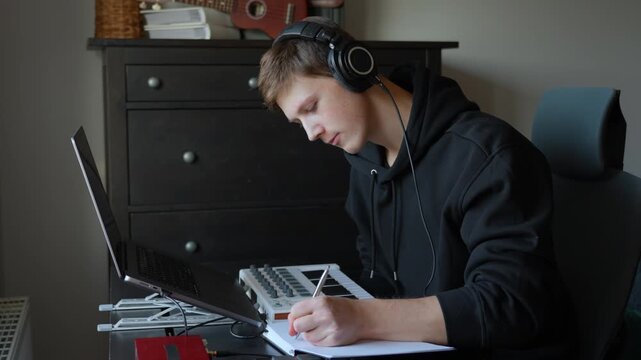 Focused songwriter wearing studio headphones pauses digital arrangement on laptop to write notes and compositional ideas in paper notebook, blending analog planning with technology in home studio