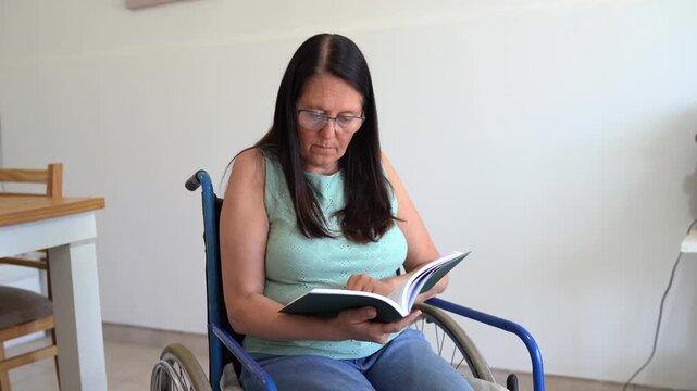 Woman With Glasses Reading Book While Sitting On Wheelchair In The House. medium shot