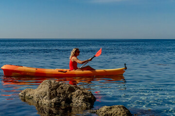 Kayaking woman ocean, blonde woman paddling orange kayak on calm blue sea, clear sky, copy space.