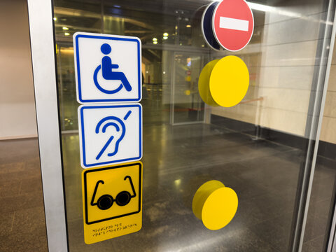 Signs for wheelchair users, deaf and blind people on glass panel near entrance with sliding doors. Sign for blindness people with Braille text and accessible area for deaf people and wheelchair users