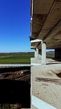 Sunlight casts shadows on a weathered concrete balcony of an unfinished building, revealing expansive green fields and distant hills under clear skies.