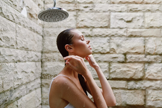 woman showering outdoors under rain showerhead with wet hair, natural stone wall background, skincare, relaxation, self-care moment in warm daylight - Powered by Adobe