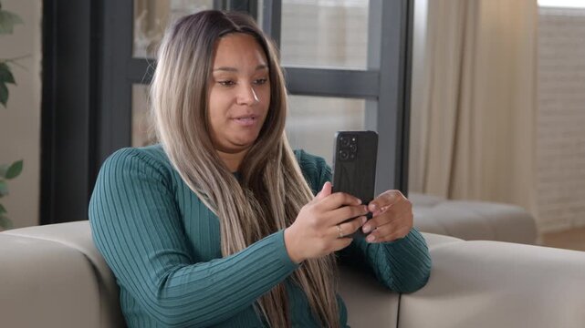 Cheerful beautiful chubby black woman relaxing on couch, making video call online using cellphone and and talking with friend about latest news while spending leisure in domestic room.