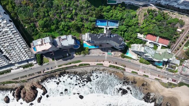 Forward top-down drone view of turquoise waves crashing on rocky coast, Vi&ntilde;a del Mar, Chile with luxury villas, pools, and coastal promenade