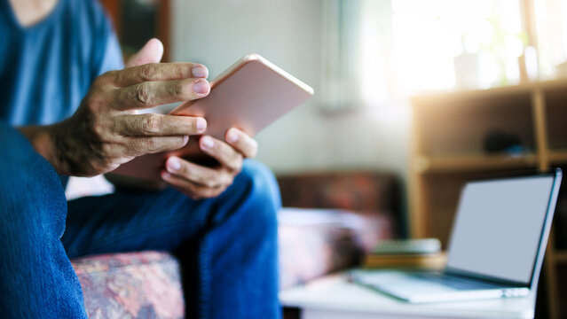 close up shot, asian man in casual clothes sitting at sofa using tablet working at home with laptop placed on the side table, online business, working at home concept