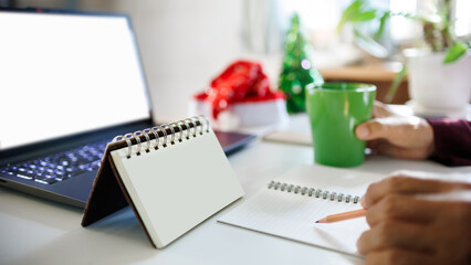 close up shot, hand holding pencil taking note on blank calendar working with laptop, red hat, christmas tree for decoration on desk, christmas holidays concept