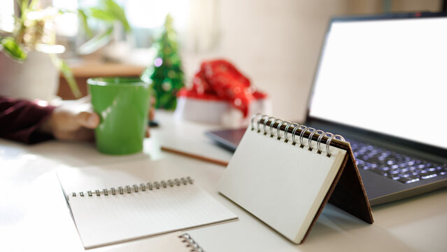 close up of calendar, notebook with computer laptop, red hat, christmas tree for decoration on desk and blurred hand holding coffee cup on background, christmas holidays concept