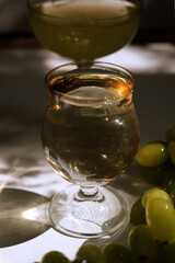Portuguese green wine Vinho Verde and bunch of green grapes in the wicker basket. Crystal glasswear. Bar counter. Sunlight and shadows on the white table, wall. The warm light of the golden hour.