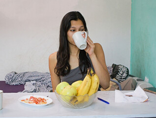Woman in a gray tank top sits at a table with fruit and a cup, drinking from a cup. Indian woman is having her breakfast. Suitable for healthy lifestyle, breakfast ideas, or morning routine topics.