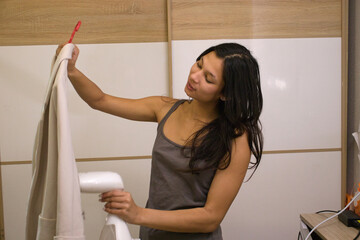 Woman in a gray tank top standing indoors uses a handheld steamer to steam a light blazer. Scene is ideal for topics like garment care, clothing tips, or daily home routines.